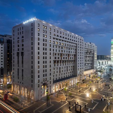 Sofitel Shahd Al Madinah Hotel Medina Exterior photo The photo shows a large modern building, likely a hotel, situated in a cityscape at dusk. The structure has a distinct architectural style with multiple stories and illuminated windows. Surrounding the building is a plaza or open area with trees and 