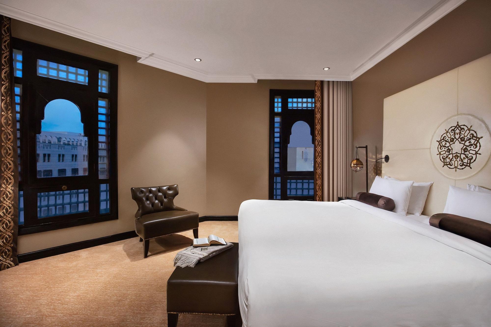 Sofitel Shahd Al Madinah Hotel Medina Exterior photo The photo shows a modern hotel room featuring a large bed with white linens, a dark upholstered headboard, and a tufted armchair in the corner. There are large windows with decorative frames that allow natural light to enter the room, and the walls a