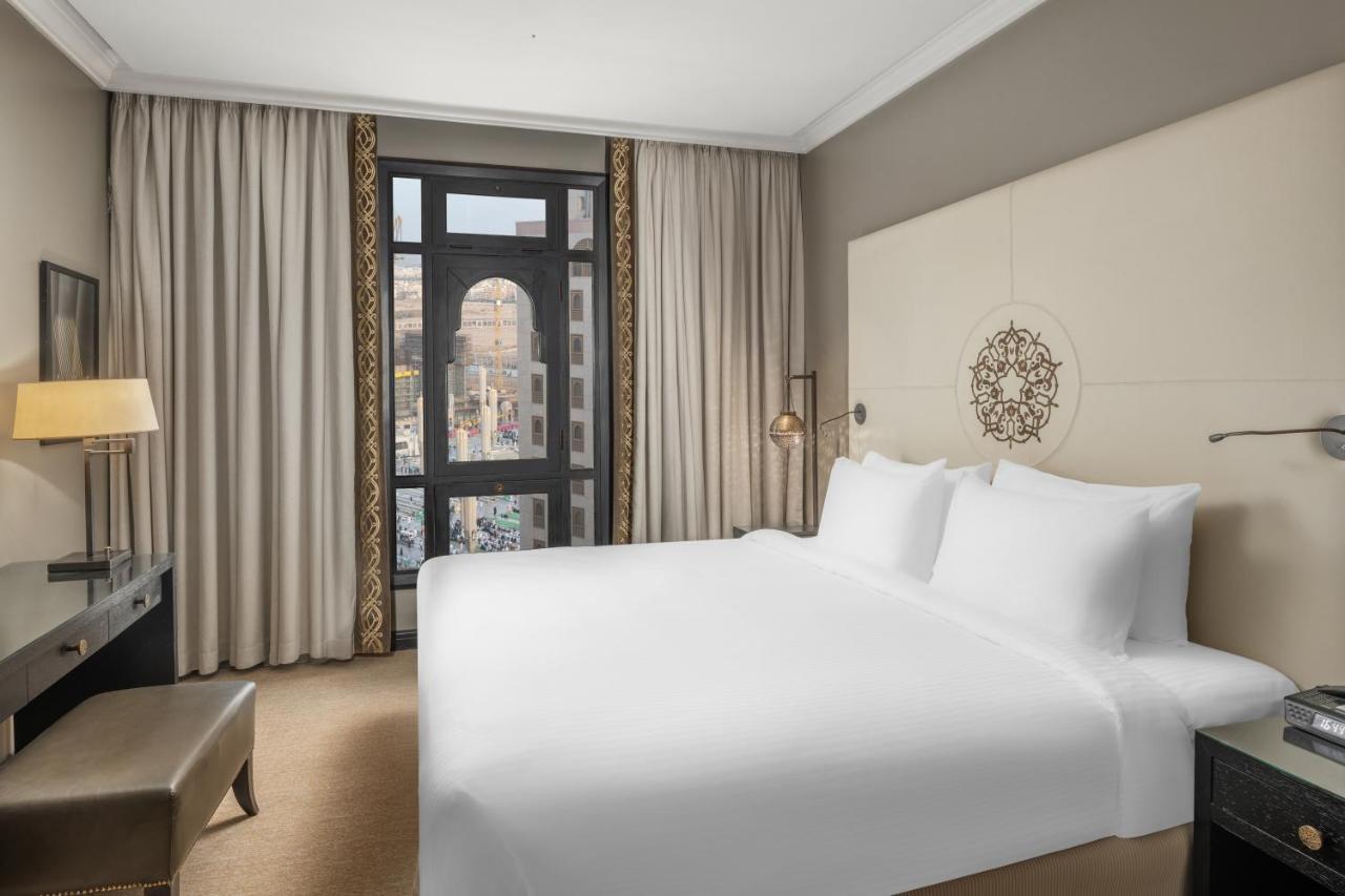 Sofitel Shahd Al Madinah Hotel Medina Exterior photo The photo shows a modern hotel room featuring a large bed with white linens. There are beige walls and plush carpet, contributing to a clean and contemporary aesthetic. Large curtains frame a window that provides a view of the outside, likely a citys