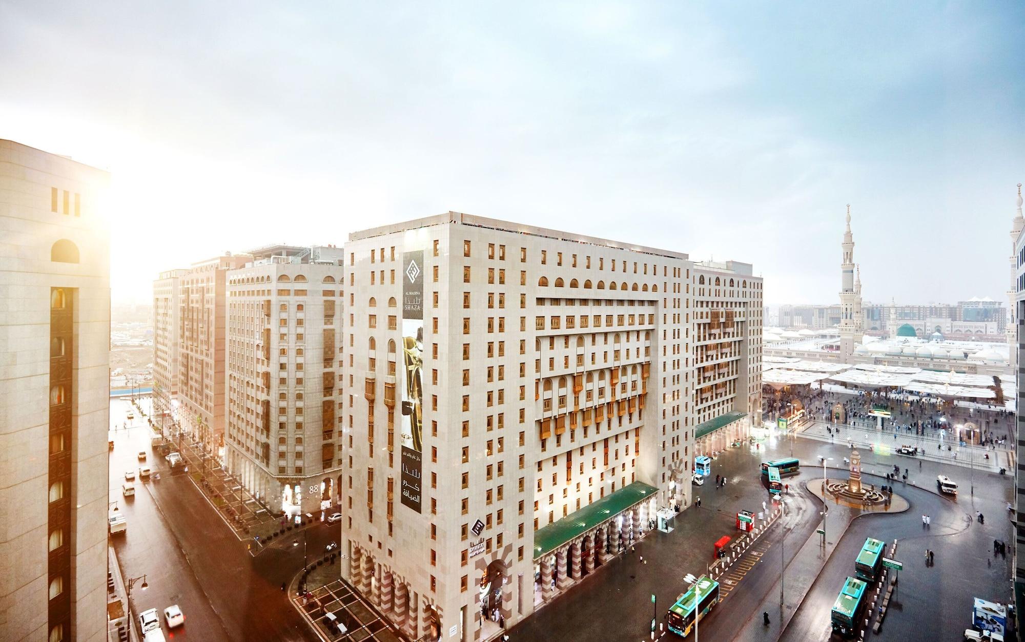 Sofitel Shahd Al Madinah Hotel Medina Exterior photo The photo shows an urban landscape featuring several large buildings, which appear to have a modern architectural style. The scene is depicted during a time of day when sunlight is visible, suggesting either early morning or late afternoon. In the fo