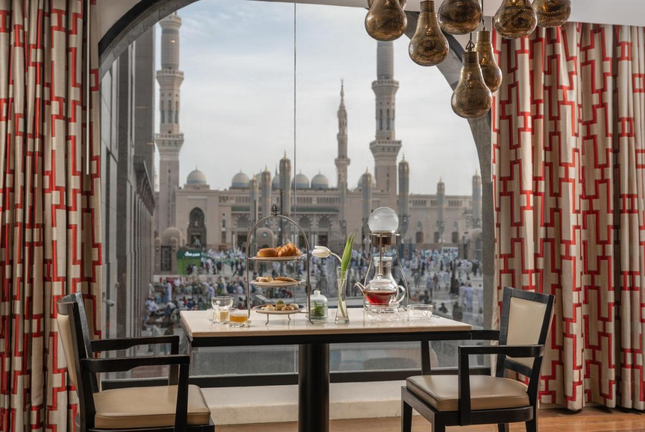 Sofitel Shahd Al Madinah Hotel Medina Exterior photo The photo shows a dining setup inside a room with a large window. A table is set for a meal, featuring a tiered stand with various foods, a teapot, and cups. The room is decorated with elegant furniture and patterned curtains. Through the window, the