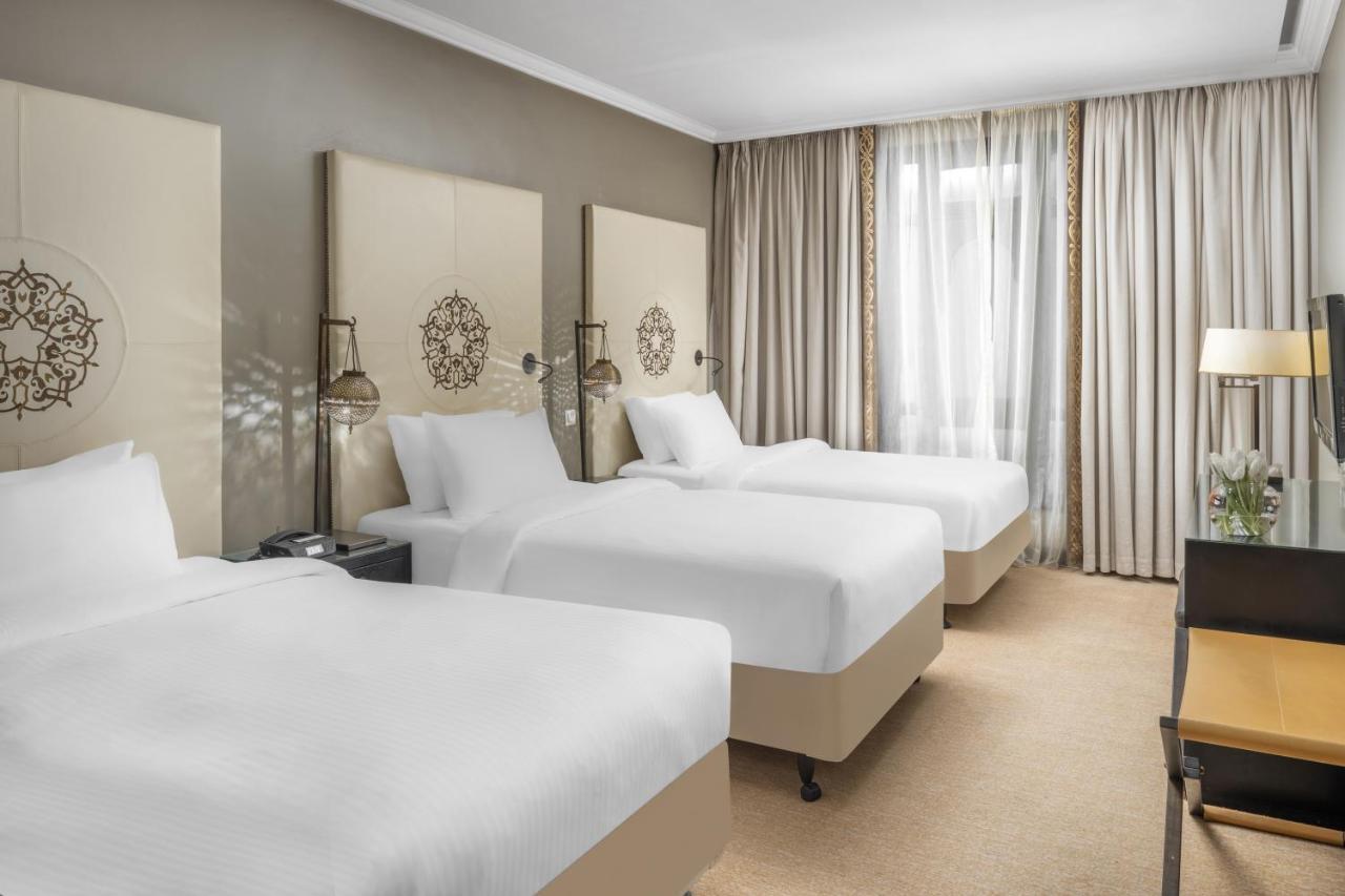 Sofitel Shahd Al Madinah Hotel Medina Exterior photo The photo shows a hotel room featuring three beds, each made with white bedding. The beds are positioned against a light-colored wall adorned with decorative headboards. There are two bedside tables with lamps next to the beds. The room is well-lit, 