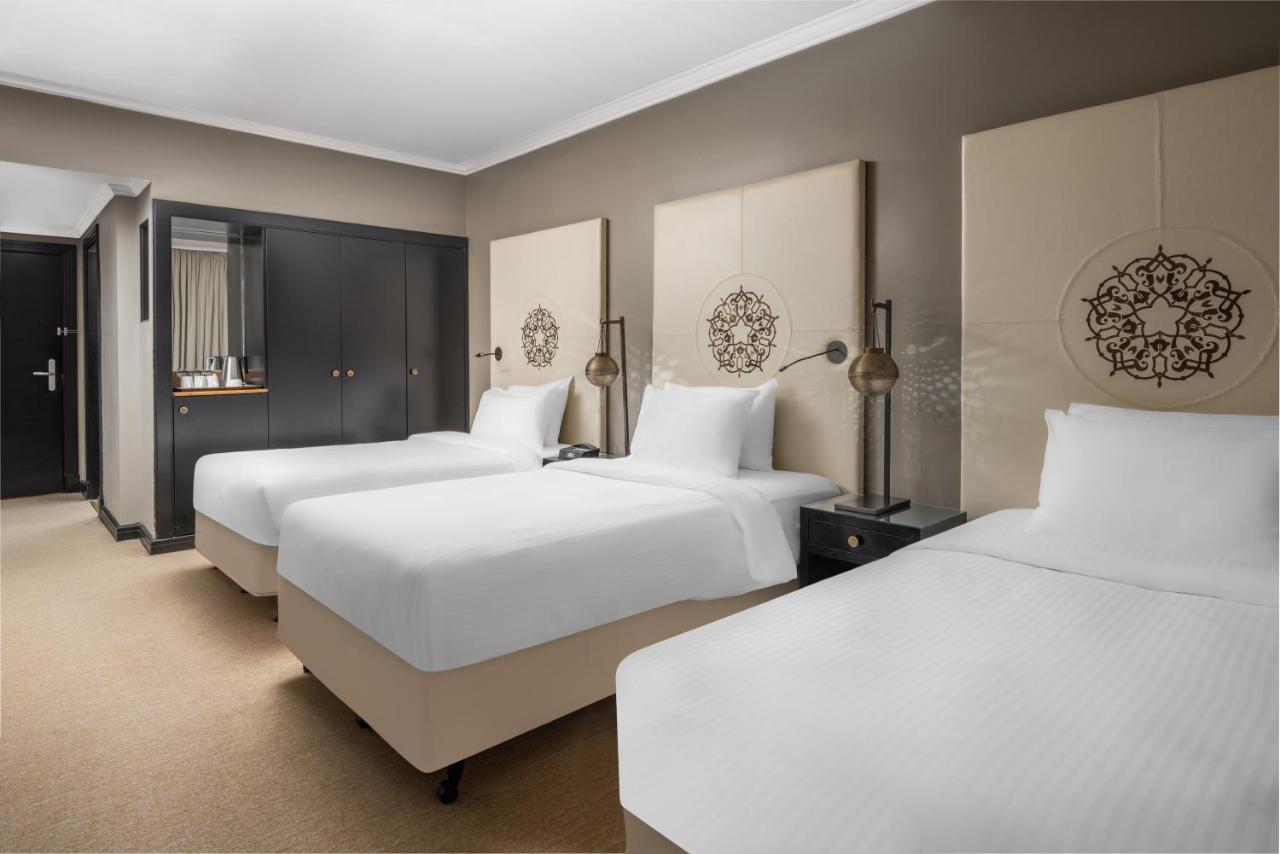 Sofitel Shahd Al Madinah Hotel Medina Exterior photo The photo shows a hotel room with three beds arranged neatly. The beds have white linens and are placed on beige bases. Behind the beds, there are decorative wall panels featuring a circular design. On either side of the beds, there are bedside table