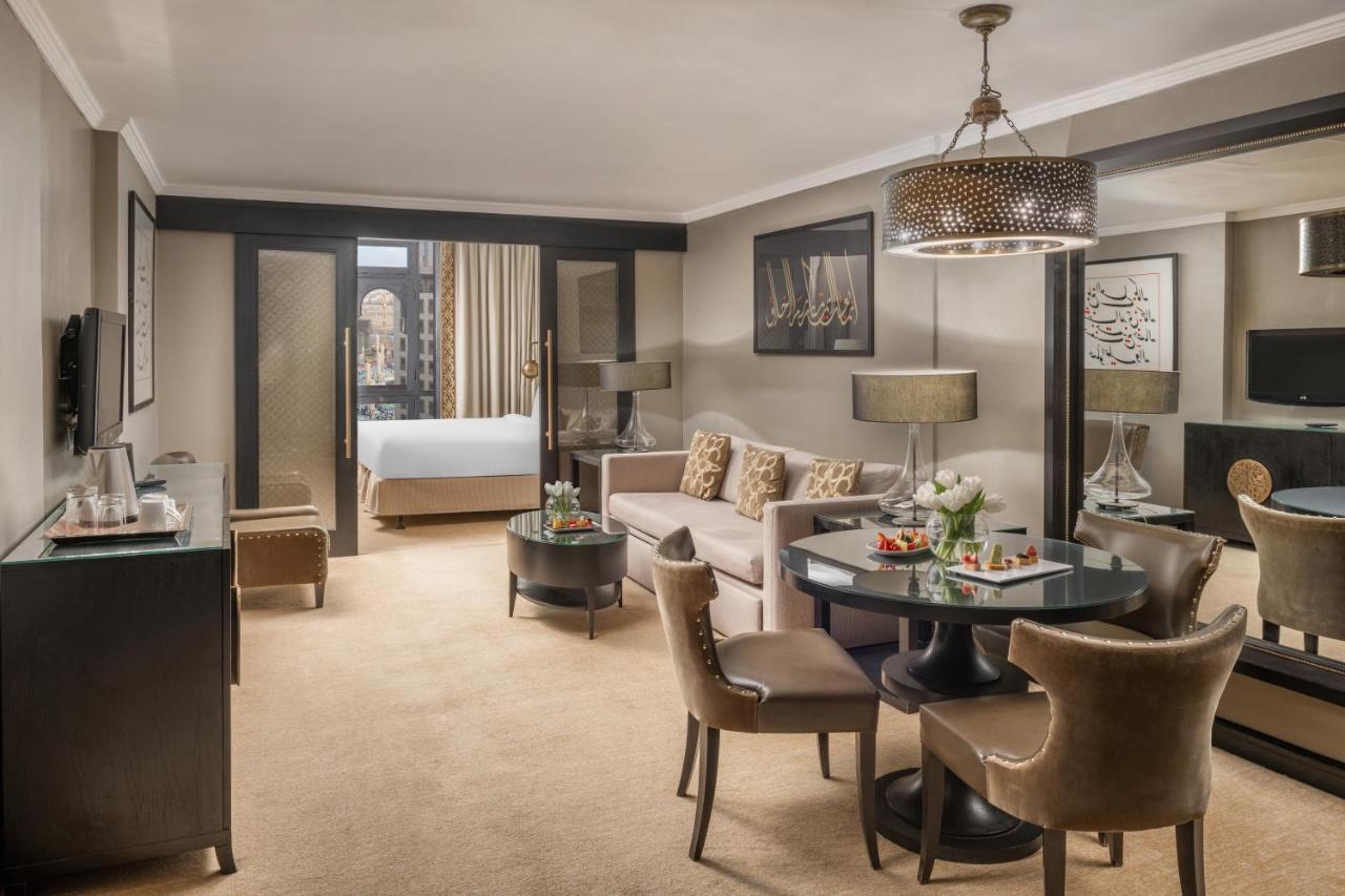 Sofitel Shahd Al Madinah Hotel Medina Exterior photo The photo shows a well-appointed hotel suite. It features a cozy living area with a beige sofa and several decorative cushions. A circular table with chairs is set in the center, likely for dining or working. There is a decorative chandelier overhead