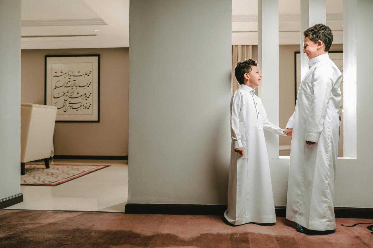 Sofitel Shahd Al Madinah Hotel Medina Exterior photo The photo shows two young boys dressed in traditional white clothing, likely thobes. They are standing in a hallway, engaged in a conversation or interaction. The setting appears to be an indoor space with modern decor, including a piece of artwork o