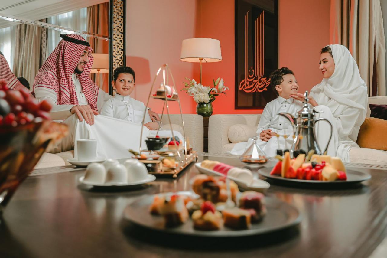 Sofitel Shahd Al Madinah Hotel Medina Exterior photo The photo depicts a cozy and inviting indoor setting where a family appears to be spending time together. In the foreground, there are plates of desserts and fruits arranged on a table, showcasing a variety of colorful treats. 

In the background, a 