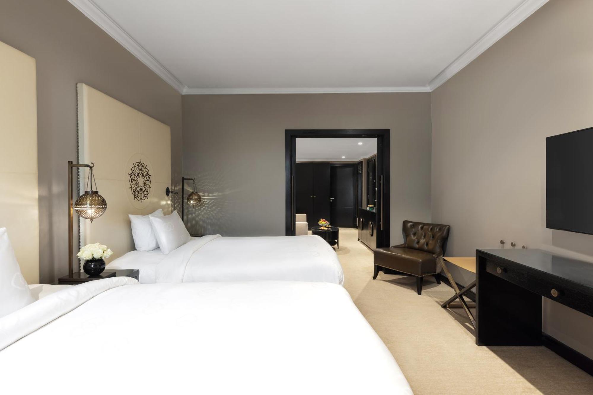Sofitel Shahd Al Madinah Hotel Medina Exterior photo The photo shows a hotel room with a modern design. It features two white beds with neatly arranged pillows, a simple beige wall with decorative accents, and stylish bedside lamps. There is a small seating area with a brown chair and a side table, as 