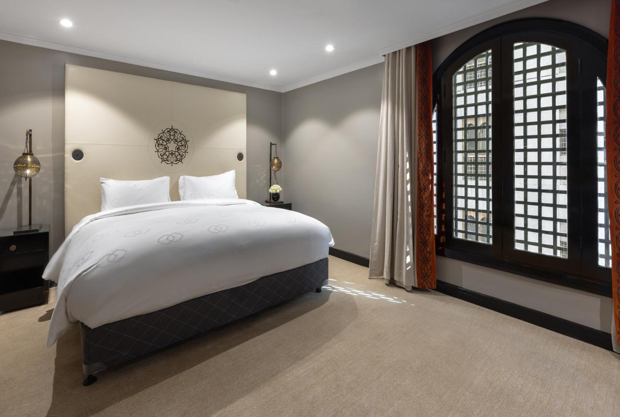 Sofitel Shahd Al Madinah Hotel Medina Exterior photo The photo shows a modern hotel bedroom. It features a large bed with white bedding and two pillows. The walls are painted in neutral tones, and there is decorative wall art above the bed. To the right, there are large windows with patterned shutters,