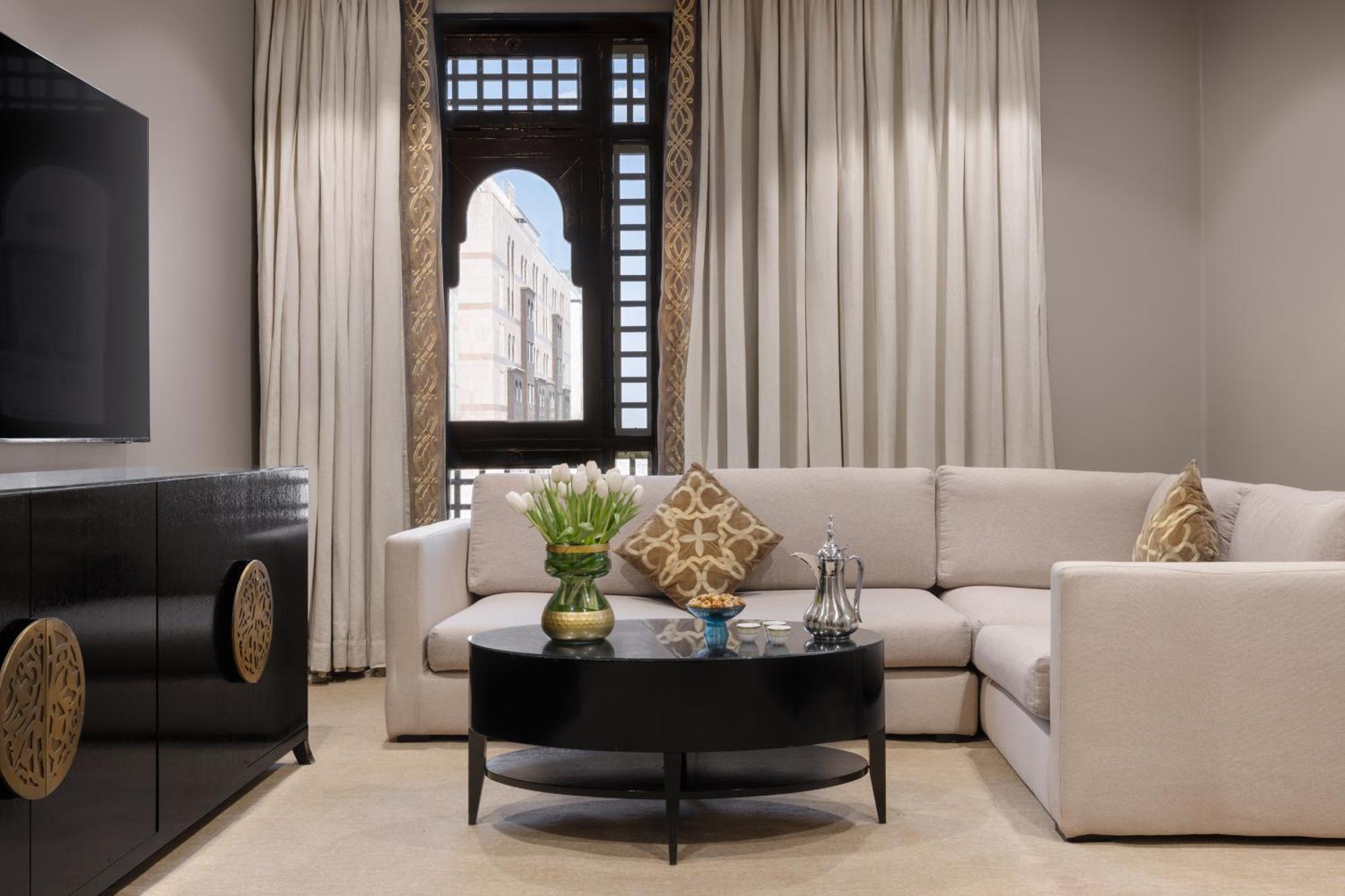 Sofitel Shahd Al Madinah Hotel Medina Exterior photo The photo shows a stylish and modern living room. It features a light-colored sectional sofa with decorative cushions, positioned around a round black coffee table. On the table, there’s a vase with flowers and some decorative items. The room has lar