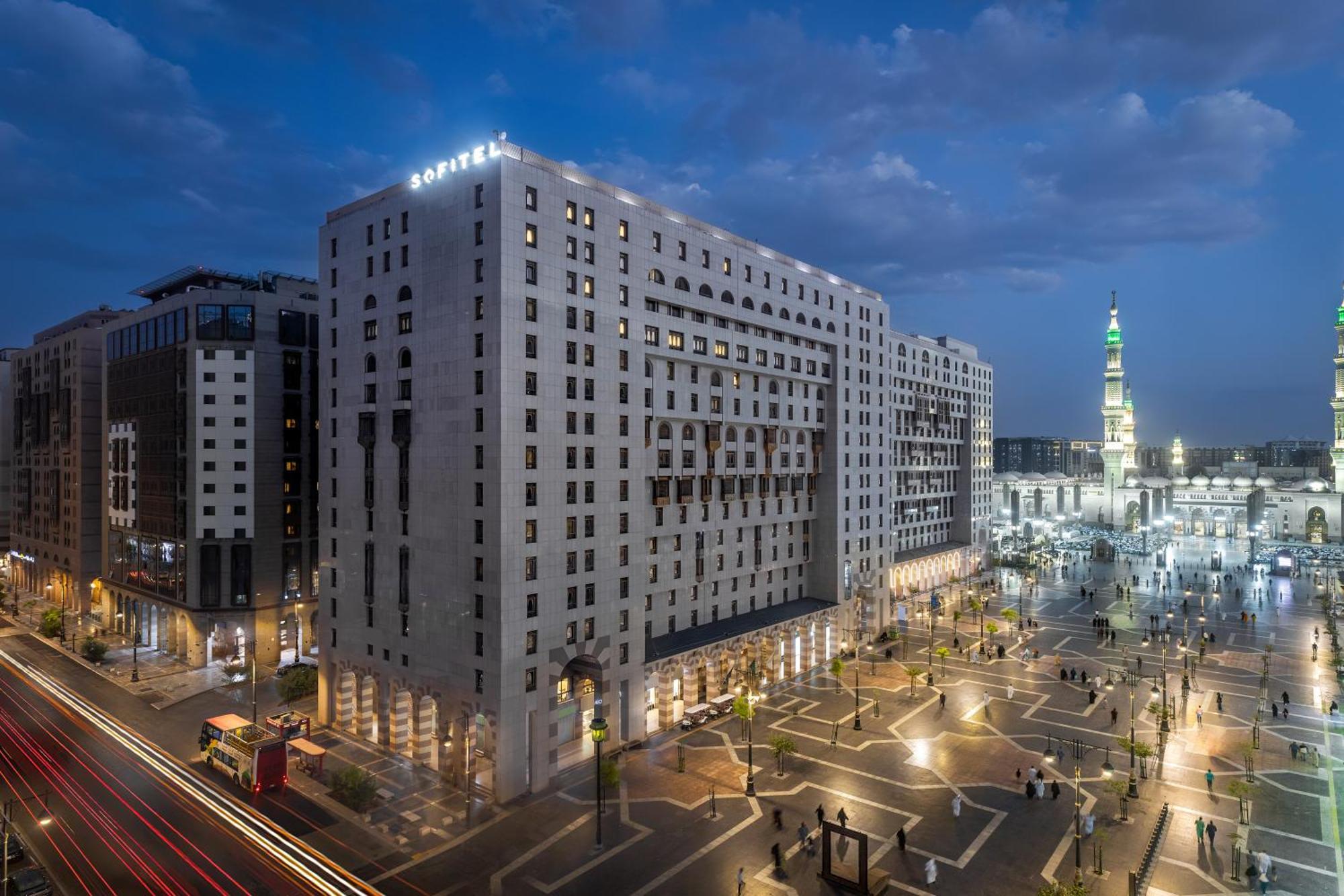 Sofitel Shahd Al Madinah Hotel Medina Exterior photo The photo shows a large modern building, likely a hotel, situated in a cityscape at dusk. The structure has a distinct architectural style with multiple stories and illuminated windows. Surrounding the building is a plaza or open area with trees and 