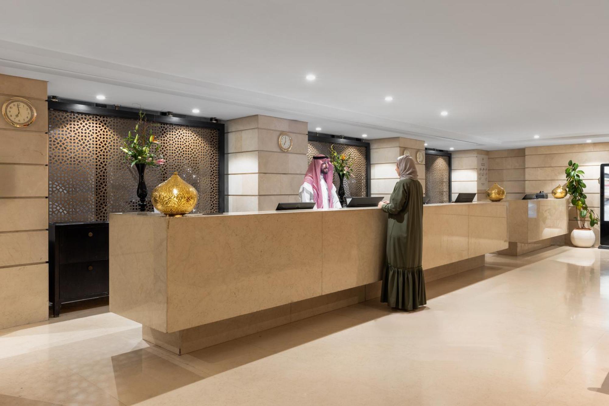 Sofitel Shahd Al Madinah Hotel Medina Exterior photo The photo shows a modern hotel reception area. There is a long, sleek reception desk made of light-colored stone. Behind the desk, a staff member is assisting a guest. The guest is wearing traditional attire. The area is well-lit with stylish lightin