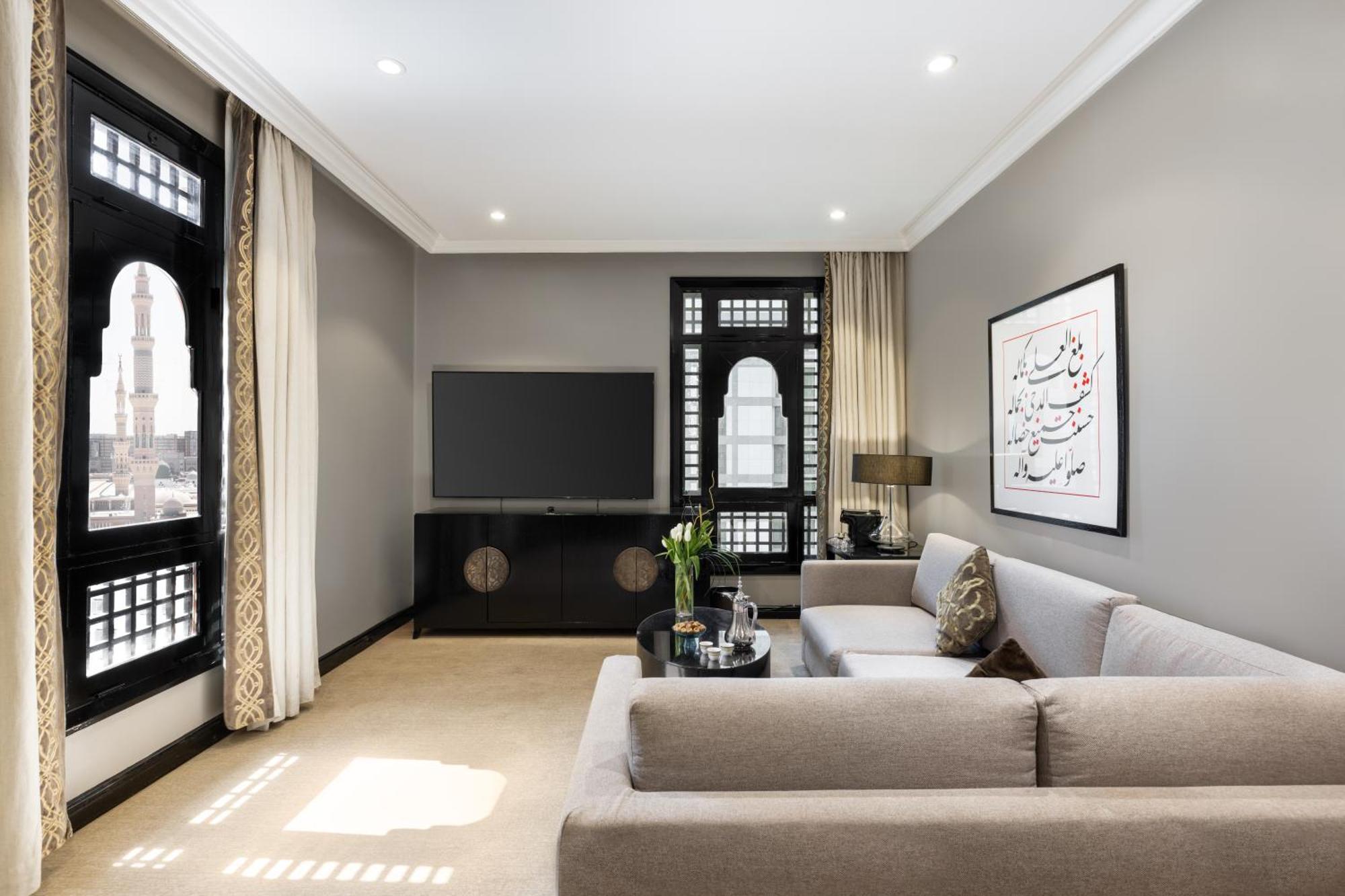 Sofitel Shahd Al Madinah Hotel Medina Exterior photo The photo depicts a modern living room with a neutral color palette. There's a comfortable light gray sofa with decorative cushions positioned toward the center. Opposite the sofa, a sleek black TV unit supports a flat-screen television. Large window