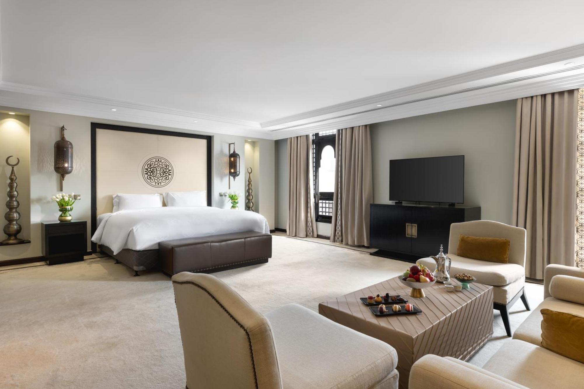 Sofitel Shahd Al Madinah Hotel Medina Exterior photo The photo shows a spacious and stylish hotel room. The room features a large bed with a dark headboard, complemented by light-colored bedding. There are two comfortable armchairs facing a coffee table, which holds some snacks and a drink. A modern te